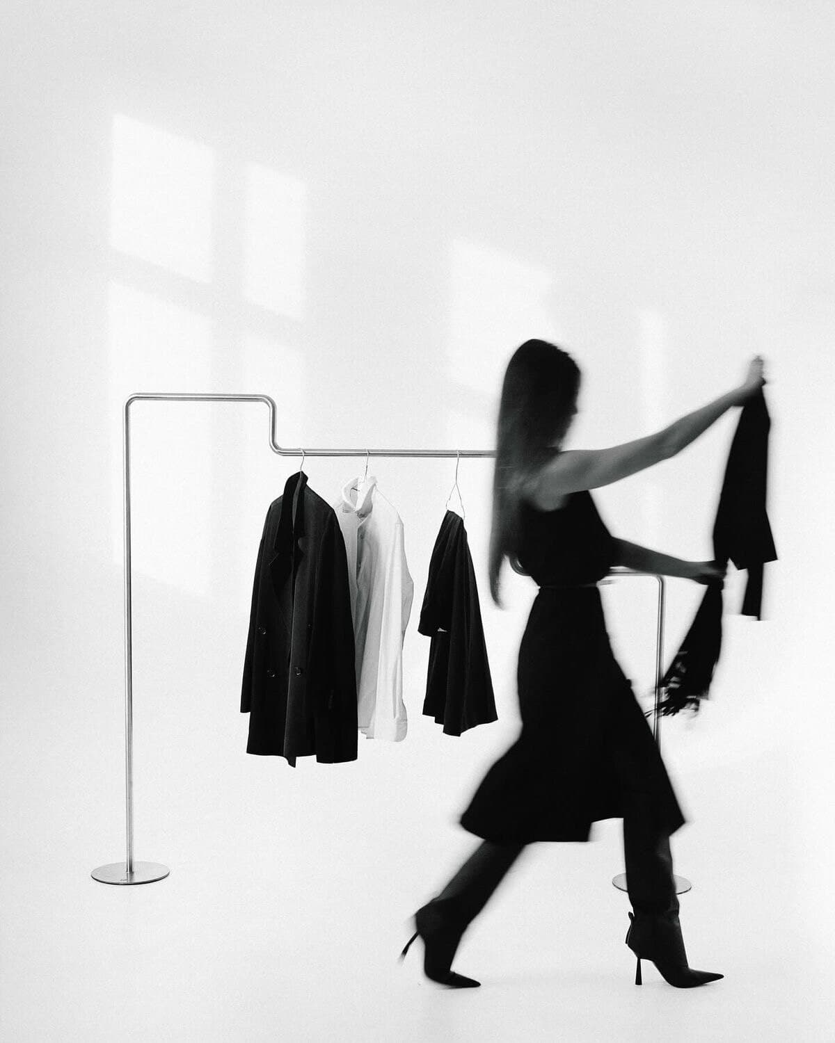 Mujer en movimiento junto a un perchero con prendas, en blanco y negro
